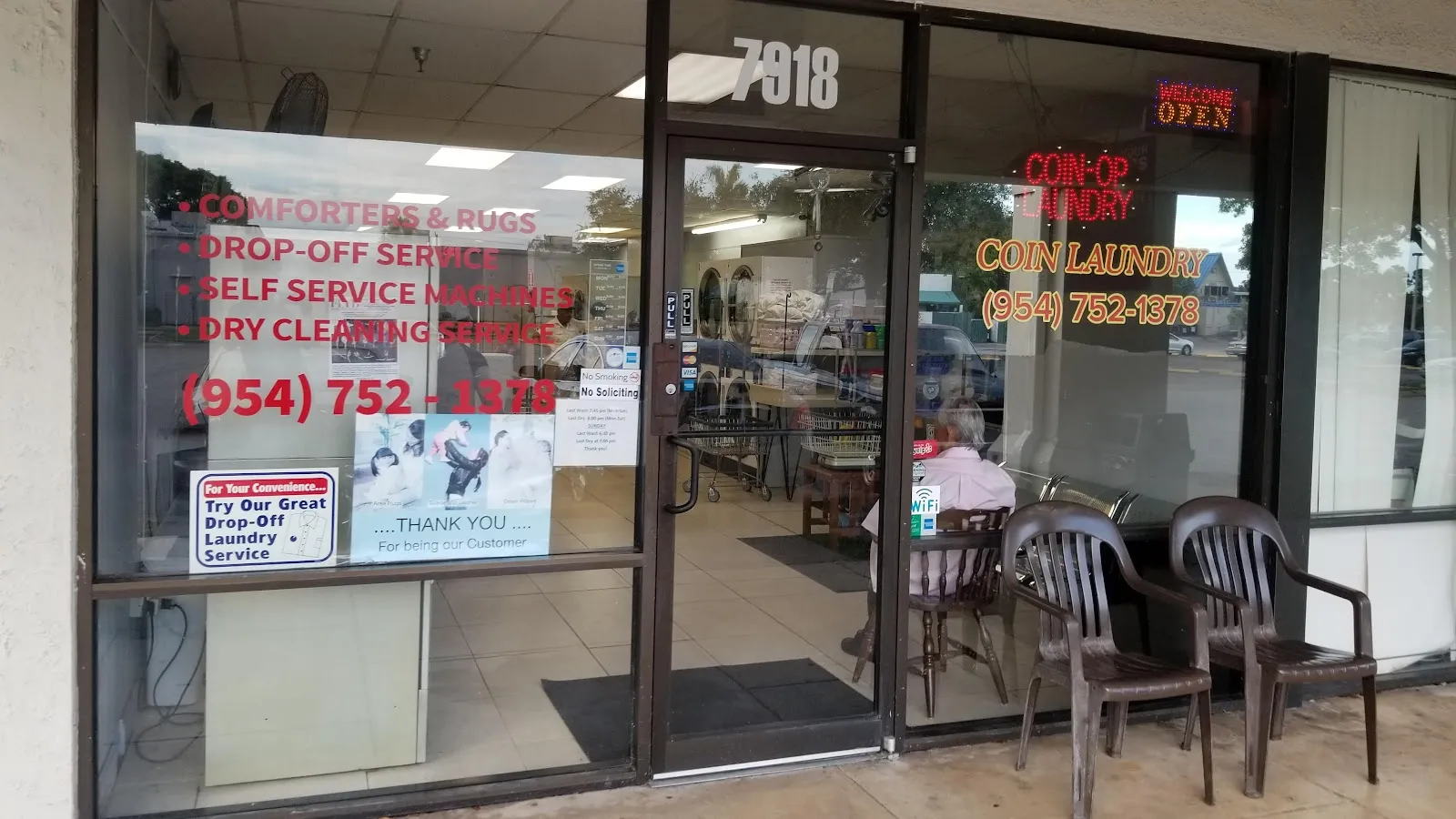 The glass entrance doors with red window lettering listing comforters and rugs, drop-off service, self service machines, and dry cleaning service, with chairs set out on the sidewalk.